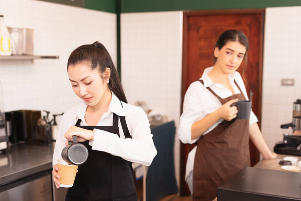 Le donne bariste asiatiche riempiono il latte nella tazza di caffè caldo da asporto mentre la donna barista caucasica la guarda per preparare il caffè per il cliente nella caffetteria. Il barista lavora al bar e al food service - Foto, immagini
