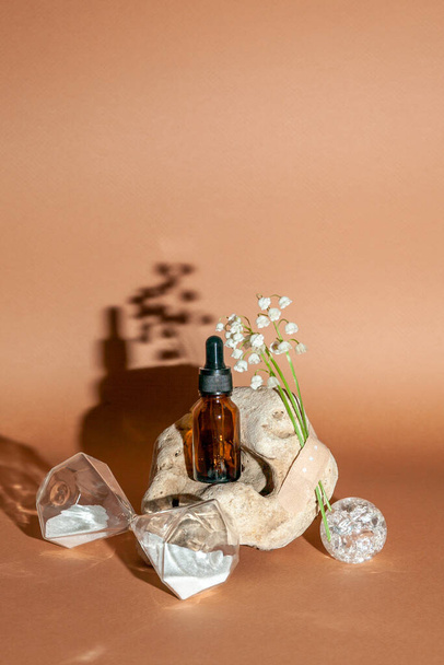 Modern still life with serum bottle on background of unusual natural stone, with bouquet of lilies of the valley and hourglass. The concept of anti-aging care products. - Valokuva, kuva