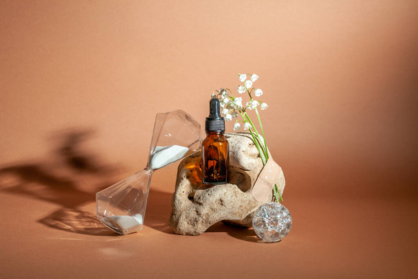 Nature morte moderne avec bouteille de sérum sur fond de pierre naturelle inhabituelle, avec bouquet de lis de la vallée et sablier. Le concept de produits de soins anti-âge. - Photo, image