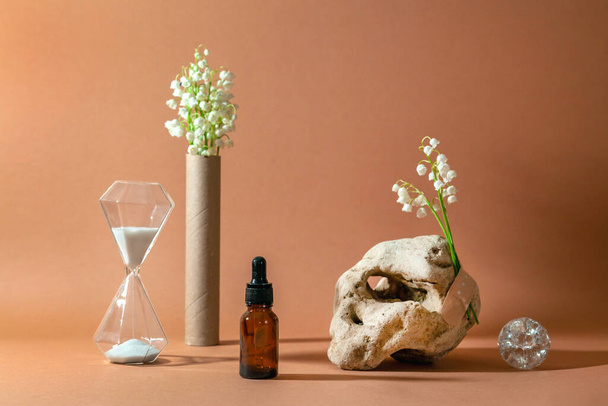 Modern still life with serum bottle on background of unusual natural stone, with bouquet of lilies of the valley and hourglass. The concept of anti-aging care products. - Photo, Image