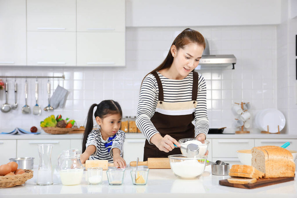 Glückliche asiatische Mutter bringt ihrer kleinen Tochter das Brotbacken in der weißen modernen Küche bei, während sie Rezept für das Kneten von Mehl zu Teig mixt - Foto, Bild