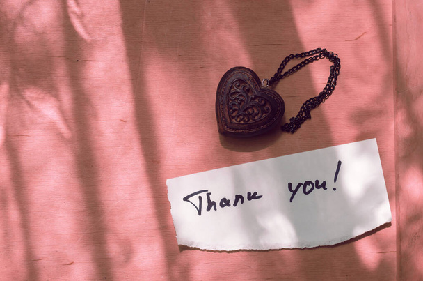 Odręczna notatka Dziękuję w białym kawałku papieru i drewnianym amulecie w kształcie serca. Tło koralowe, cienie. Miłość, koncepcja związku. Widok z góry - Zdjęcie, obraz
