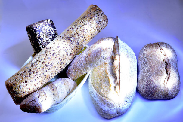 Μπαγκέτες διαφόρων σχημάτων, ψημένες από αλεύρι σιταριού και σίκαλης, βρίσκονται στο καλάθι του ψωμιού, και υπάρχουν ρολά στην αριστερή πλευρά του καλαθιού. - Φωτογραφία, εικόνα