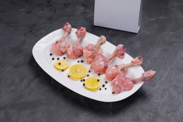Roher Hühnerlutscher, sechs Stück Hühnerlutscher auf einem weißen Teller angeordnet und mit Zitronenscheiben und schwarzem Pfeffer mit Lieferbox auf grauem texturiertem Hintergrund garniert - Foto, Bild