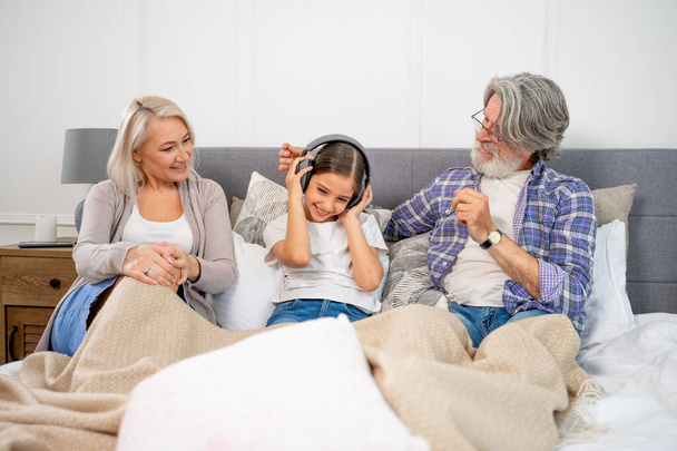 Cute little girl listening music in headphones with loving grandparents - Photo, Image