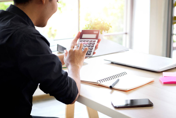 Бизнесмен или бухгалтер азиатский молодой человек держит калькулятор для расчета финансовых данных, затрат и бюджета на рабочем столе дома, бухгалтерской статистики и кредитного аналитика для оплаты.  - Фото, изображение