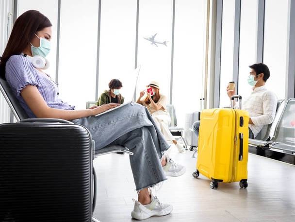 Yüz maskesi takan Asyalı genç turistler Coronavirüs enfeksiyonunu önlemek için birbirlerinden uzak oturmaya devam ediyorlar, havaalanı terminalinde dizüstü bilgisayar ve telefon kullanıyorlar, boş koltukta kırmızı haç gösterileri, sosyal mesafeler ve yeni normal seyahat  - Fotoğraf, Görsel