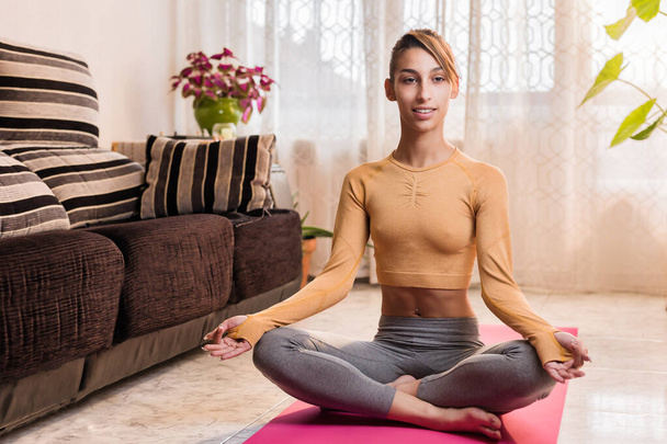 Yoga evde. Genç İspanyol kadın karantina sırasında oturma odasında sukhasana eğitimi yapıyor. Sağlık koçu egzersiz meditasyonu - Fotoğraf, Görsel