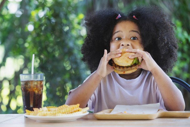 infanzia e mangiare concetto - piccola afro-americana ricci ragazza dei capelli godendo di un hamburger e patatine fritte sul tavolo.. - Foto, immagini