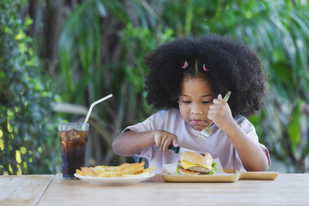 παιδική ηλικία και διατροφική έννοια - λίγο αφροαμερικανός σγουρά μαλλιά κορίτσι κρατώντας μαχαίρι και κοιτάζοντας burger και τηγανητές πατάτες στο τραπέζι. απολαμβάνοντας ανθυγιεινά τρόφιμα. - Φωτογραφία, εικόνα