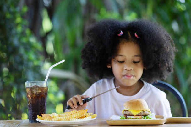 детство и концепция питания - маленькая африканская кудрявая американка с ножом в руках и глядя на бургер и картошку фри на столе.. - Фото, изображение