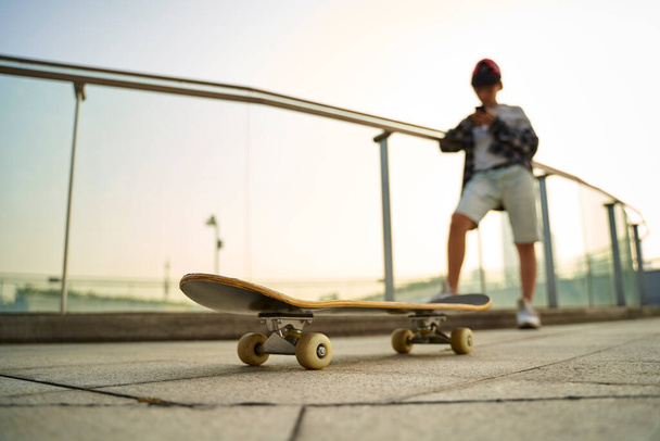 teenage asian skateboarder boy looking at mobile phone while resting, focus on the skateboard - Photo, Image