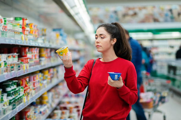 Valkoihoinen nuori kaunis nainen valitsee kahden jogurtin väliltä. Käsite kulutustottumukset ja ostokset. - Valokuva, kuva