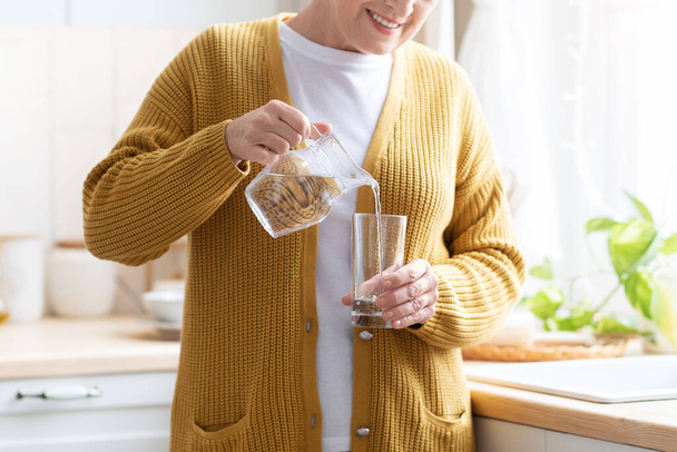 Unrecognizable grandmother pouring water into glass at home - Photo, Image