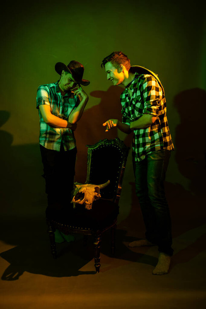 Two guys in plaid shirts and hats stand by a chair with a bull skull illuminated by multicolored light - Foto, imagen
