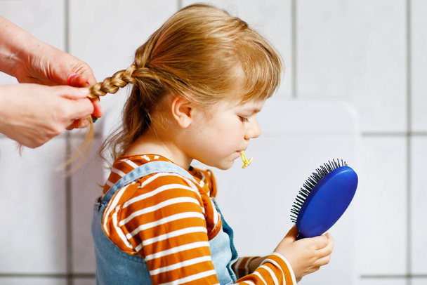 Close up photo of happy smiling toddler girl with blond hair and hands of mum brushing hairs and make plait or pigtail. Mother prepares child for kindergarten or daycare. Family morning routine. - Photo, Image