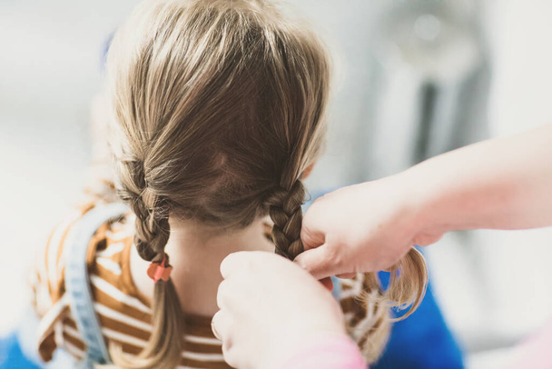 Close up photo of head of toddler girl with blond hair and hands of mum brushing hairs and make plait or pigtail. Mother prepares child for kindergarten or daycare. Family morning routine at bathroom. - Valokuva, kuva