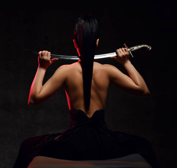 Back view of young pretty brunette woman warrior sitting topless back to camera holding sword on shoulders - Fotoğraf, Görsel