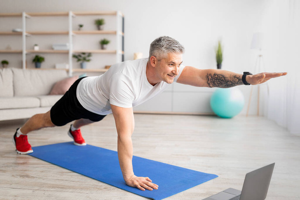 Online domestic sports. Senior man exercising to video on laptop computer, doing plank and raising one arm - Photo, Image