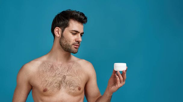 Portrait of naked model guy holding white jar with effective cream for face, posing isolated over blue background - Фото, изображение