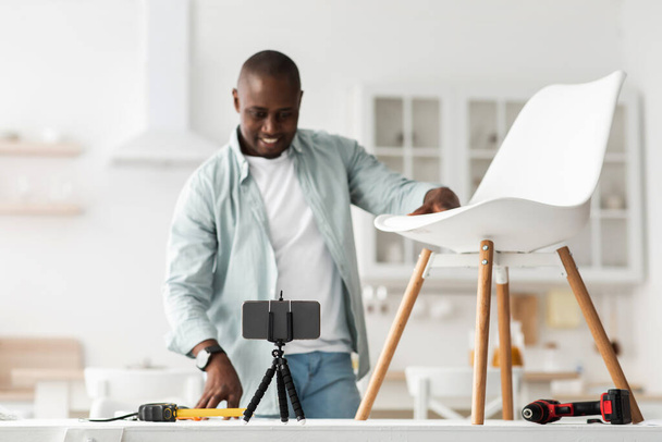 Blogger moderno. Feliz hombre negro mostrando su silla ya hecha y mirando a la cámara del teléfono inteligente, grabación de vídeo - Foto, imagen