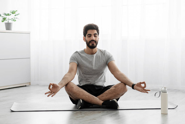Cheerful arabian bearded millennial man sits on mat in lotus position, rest on floor with bottle of water - Фото, изображение