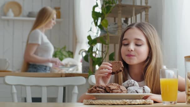 Niña adolescente niña niña colegiala sentada en la mesa en la cocina casera comiendo deliciosas galletas de chocolate caseras disfrutando del desayuno en el fondo de la madre borrosa lavar platos plato - Metraje, vídeo