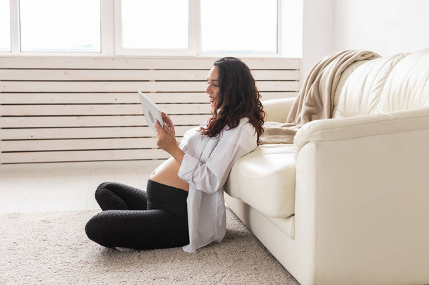 Schwangere mit Tablette auf Teppich neben Couch im heimischen Wohnzimmer - Foto, Bild