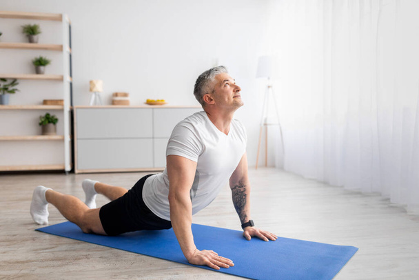 Actieve senior man doet cobra pose op yoga mat, oefenen in de woonkamer interieur, lege ruimte - Foto, afbeelding