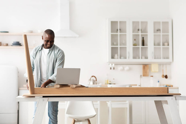 Furnishing house. Happy african american man assembling wooden table and looking at online instructions on laptop - Zdjęcie, obraz