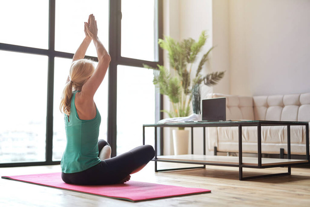 Sportos érett szőke nő gyakorló jóga, ül lótuszpozícióban a padlón, és néz online edzés segítségével laptop otthon - Fotó, kép