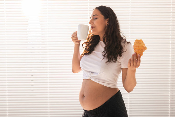 Glückliche Schwangere mit Croissant-Brötchen zu Hause. Schwangerschaft, Essen und Menschen-Konzept - Foto, Bild