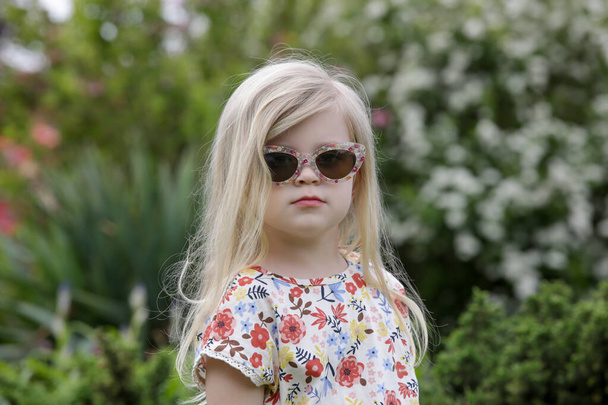 Portrait of fashionable toddler girl with long blonde hair in retro sunglasses and floral dress on a sunny summer day in park. - Фото, изображение