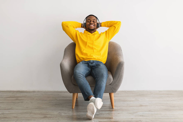 Chico negro sentado escuchando música en auriculares, fondo gris - Foto, imagen