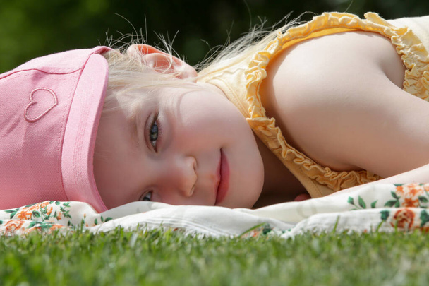 Portret uroczej dziewczynki w różowym letnim kapeluszu leżącej na zielonej łące - Zdjęcie, obraz