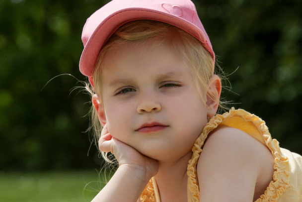Porträt eines entzückenden Kleinkindes mit rosa Sommermütze, das sich auf eine grüne Wiese legt - Foto, Bild