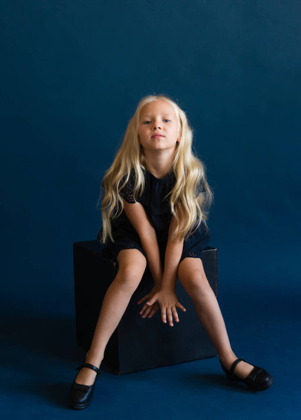 beautiful blonde in a dark blue dress on a solid blue background. The photo is great for demonstrating the character of a child - daydreaming, enthusiasm, vulnerability, touching, beauty, stubbornness, harm, cunning, coquetry. - Fotó, kép
