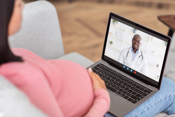 Medizinische Online-Beratung. Werdende Frau hat Videoanruf mit freundlichem schwarzen Arzt auf Laptop von zu Hause aus - Foto, Bild
