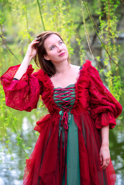 beautiful brunette young woman in red gown standing by lake under tree - Valokuva, kuva