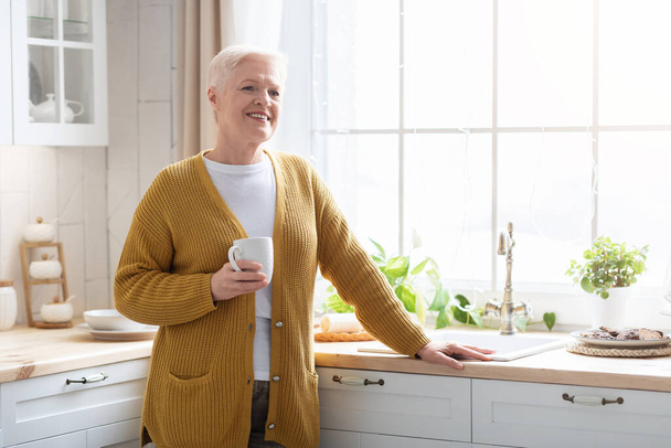 Smiling attractive senior lady with mug, drinking coffee at kitchen - Фото, изображение