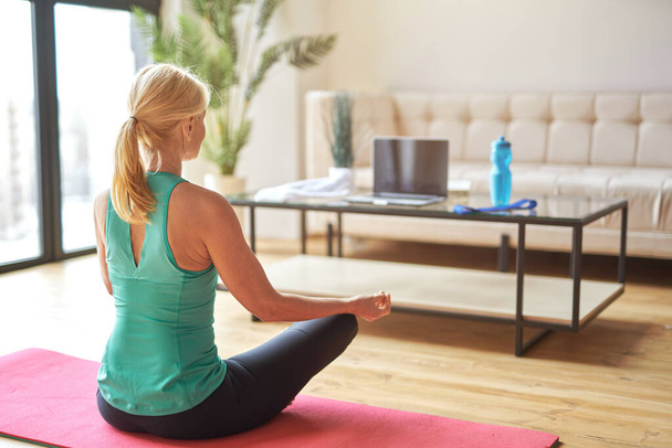 Vista posterior de la mujer rubia madura deportiva sentada en posición de loto en el suelo y haciendo ejercicios de respiración en casa durante el día - Foto, Imagen