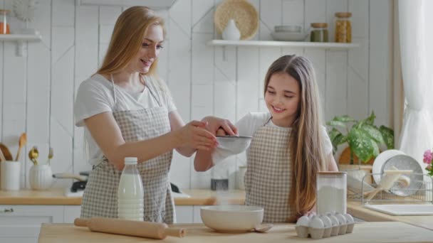 Aikuinen äiti blondi nainen ja pieni tytär teini-ikäinen tyttö käyttää esiliinat oppia kokki yhdessä pelkuri vehnä valkoinen jauhot rauta seula valmistella ruokalaji taikina resepti lisätä ainesosa seisoo keittiössä - Materiaali, video