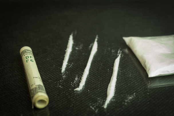 Drugs and dollars on glass, black table. Narcotic powder divided into stripes on a mirrored table, one hundred dollar tube for drug use - Photo, Image