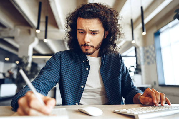Handsome curly guy is making notes at the working table in open space office - Foto, imagen