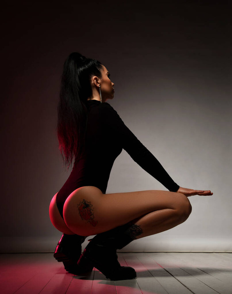 Young sexy slim brunette woman in black thongs bodysuit sits squatting back to camera showing tattoo on buttocks - Fotó, kép