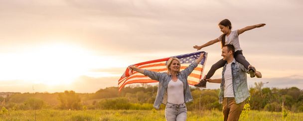Young parents with their daughter holding American flag in countryside at sunset. Independence Day celebration - Photo, Image