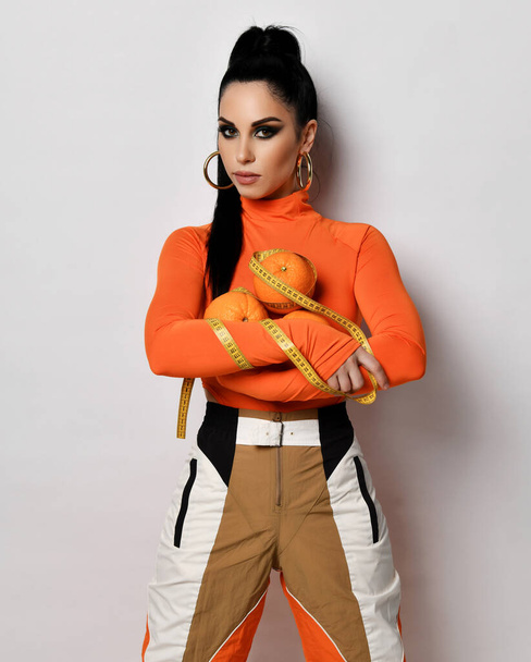 Young slim brunette woman in orange sportswear pants and turtleneck stands holding fresh oranges and measuring tape - Photo, Image