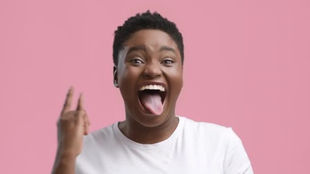 Lustige Afrikanerin gestikuliert auf Felsschild mit Zunge, rosa Hintergrund - Filmmaterial, Video