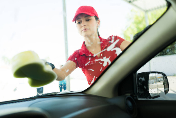 勤勉な女性労働者は車のフロントガラスをきれいに取得します。車の中で若い女性と従業員は車を洗う  - 写真・画像