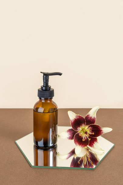 Brown glass bottle with pump of cosmetic products and lily flower on mirror, beige background. Natural Organic Spa Beauty Cosmetic concept Mockup. - Foto, Imagem